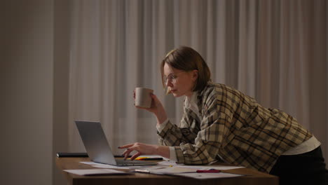 A-young-beautiful-business-woman-works-from-home-with-a-cup-of-coffee-and-looks-into-a-laptop-screen-standing-at-the-table.-View-mail-and-drink-coffee.-Remote-work-from-home